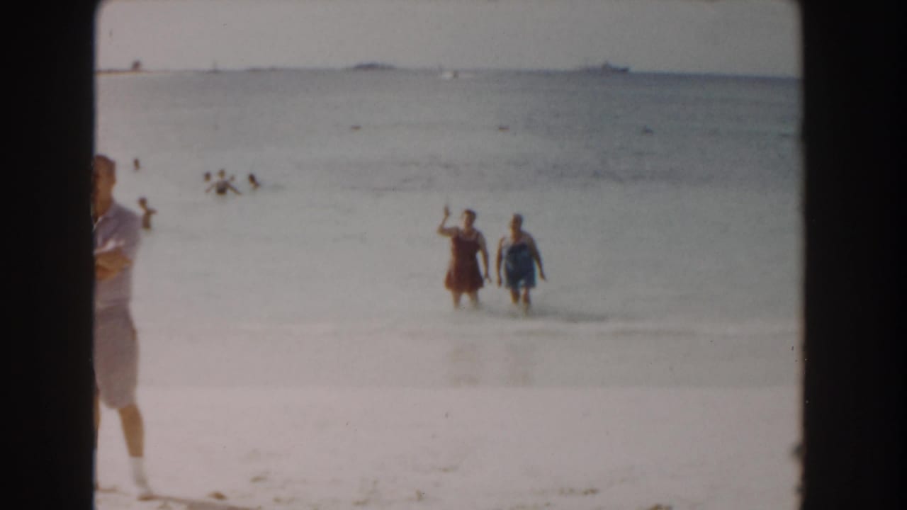 Two women stand in shallow water while a beachgoer is partly visible at the left edge and swimmers are offshore.