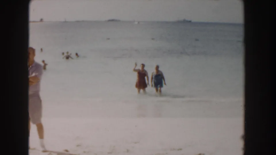 Two women in swimsuits wade in the shallow Nassau surf, with a partial beachgoer at the frame edge and distant swimmers beyond.