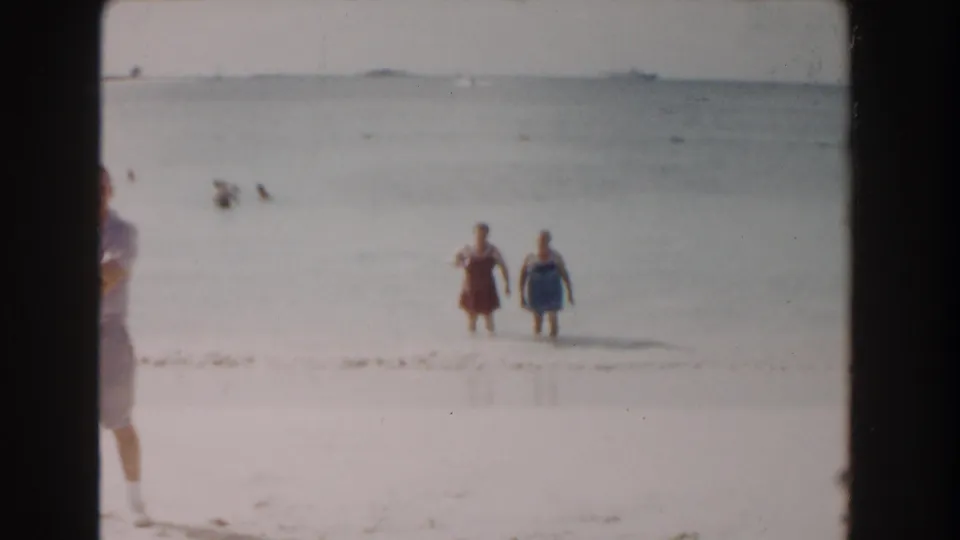 The 1961 Nassau beach scene shows two women moving toward shore through shallow surf, with bathers in the distance and a cropped figure at left.