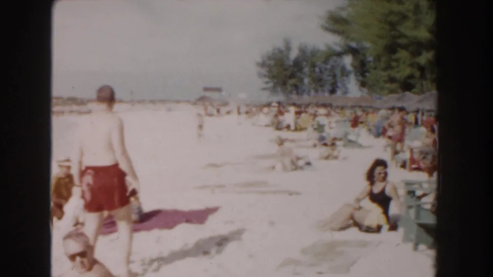 A wider Nassau beach view shows a man in red swim trunks walking past seated bathers, umbrellas, trees, and crowded sand in 1961.