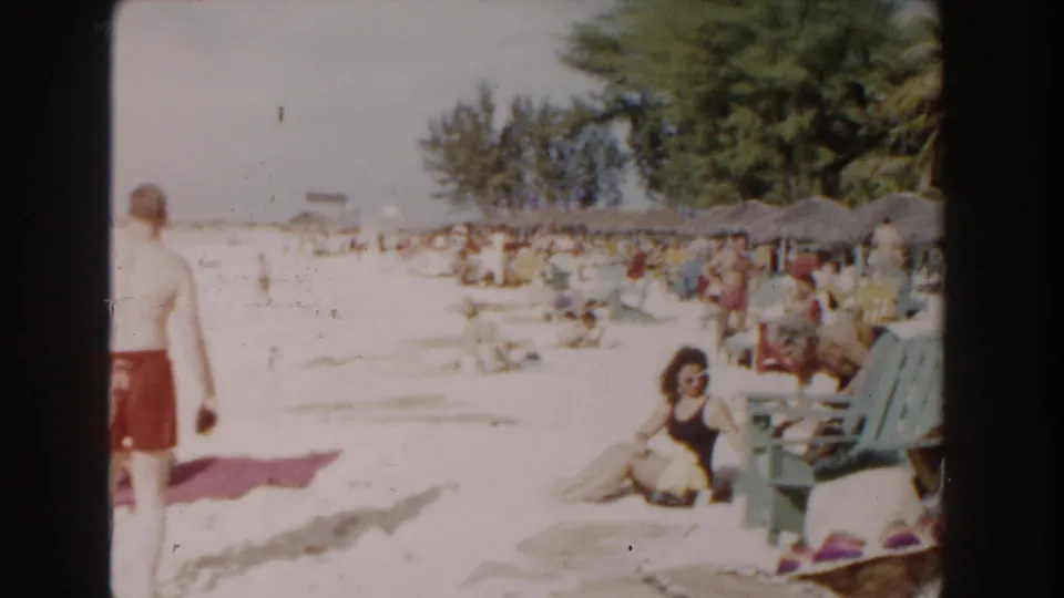 Nassau beach scene from 1961 with people on white sand, colorful chairs, and shaded thatched umbrellas in soft 8mm color.
