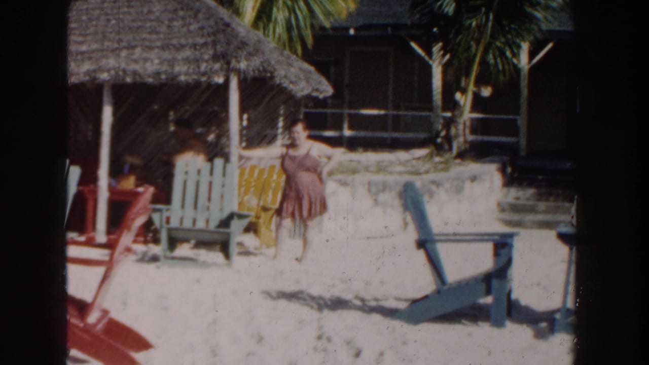 Woman in a brown dress stands by colorful beach chairs near a thatched shelter.