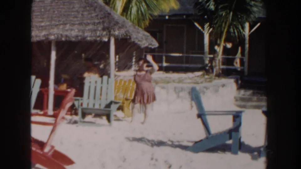 In this 1961 Nassau beach frame, a woman in a brown dress waves toward the camera beside colorful chairs and a thatched shelter.