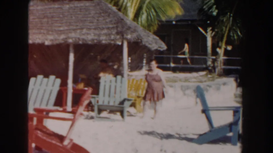 Nassau beach view with a woman in a brown dress, Adirondack-style chairs, palm trees, and a shaded thatched shelter in faded 8mm color.