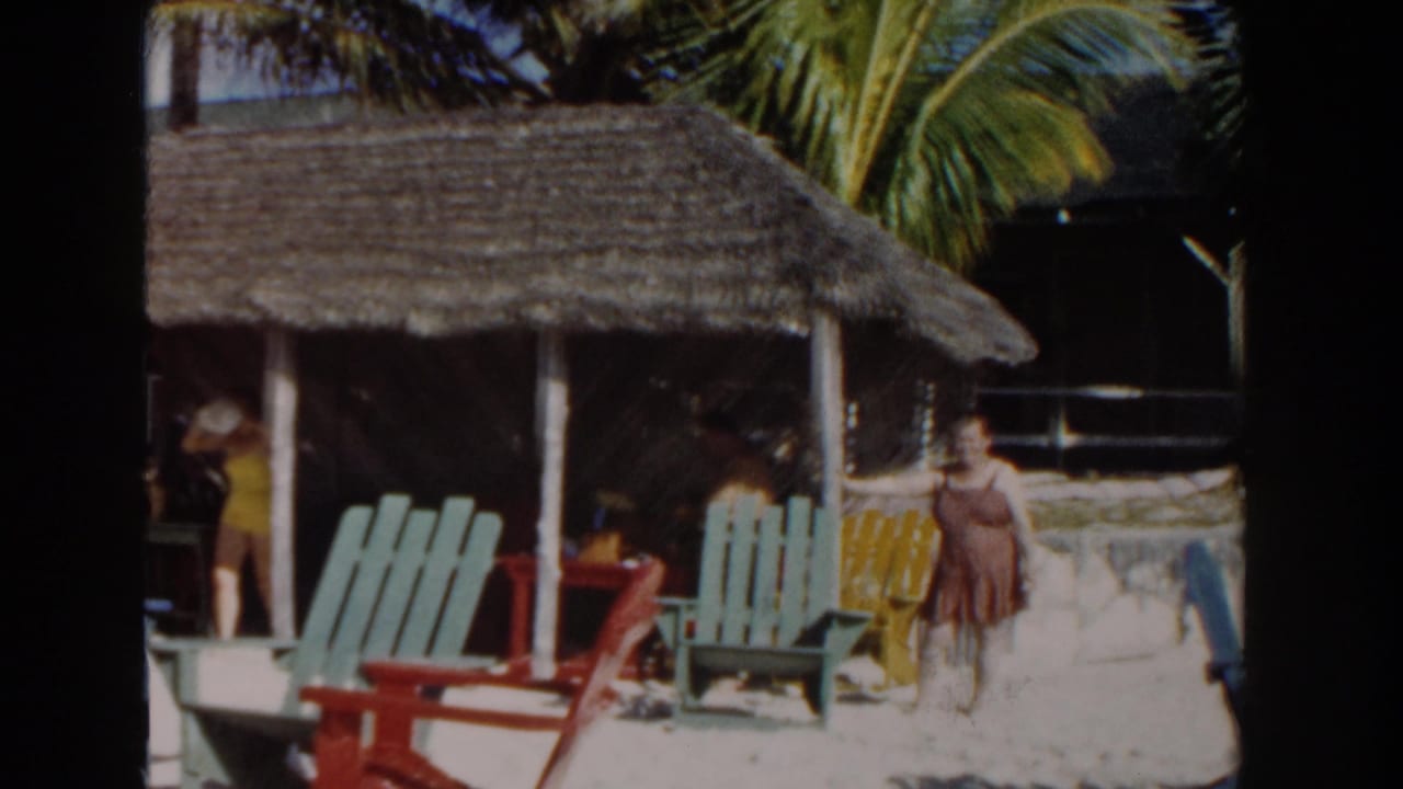 Woman in yellow walks near a thatched beach hut with colorful chairs in front.