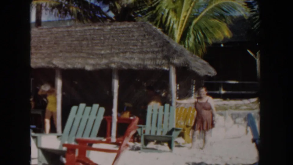 1961 Nassau resort scene with a woman in yellow near a thatched hut, palm trees, sandy ground, and red and green chairs.
