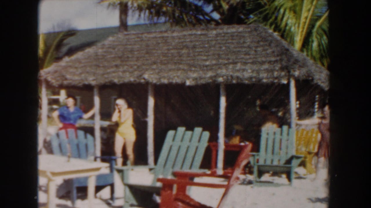 Two women stand by a thatched hut with green and red chairs on sandy ground.