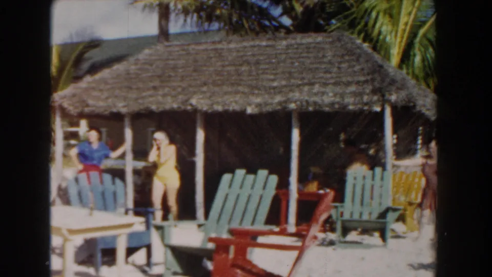 At a Nassau beach resort in 1961, two women stand near a thatched cabana with colorful chairs and palm trees nearby.