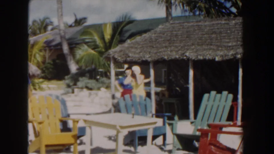 1961 Nassau home-movie frame showing a woman in yellow near a thatched hut, with bright chairs, palms, and sandy resort grounds.