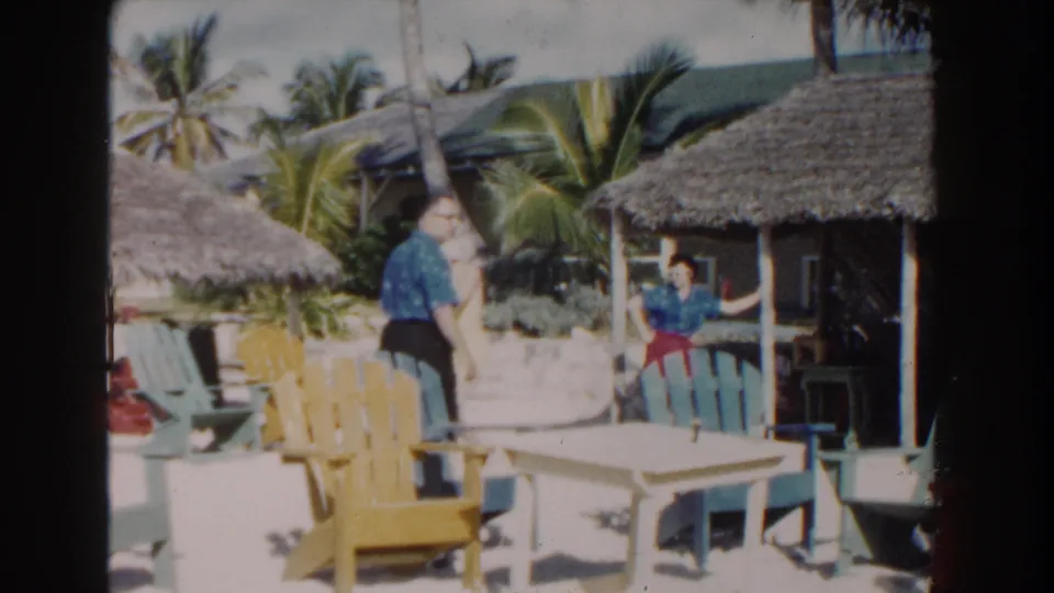 A 1961 Nassau resort view shows a woman in a blue top and red pants near a palm, thatched hut, and colorful sandy beach seating.