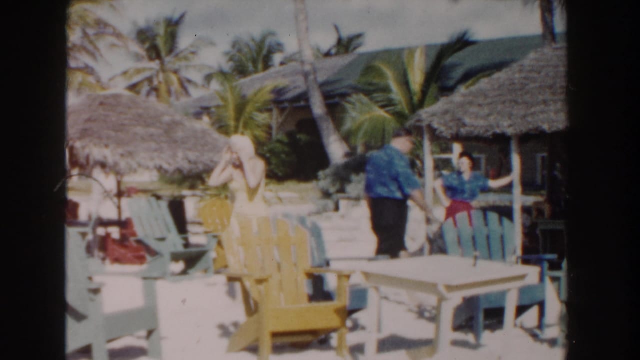 Nassau beach scene with colorful chairs, palm trees, thatched cabanas, and vacationers in early-1960s 8mm footage.