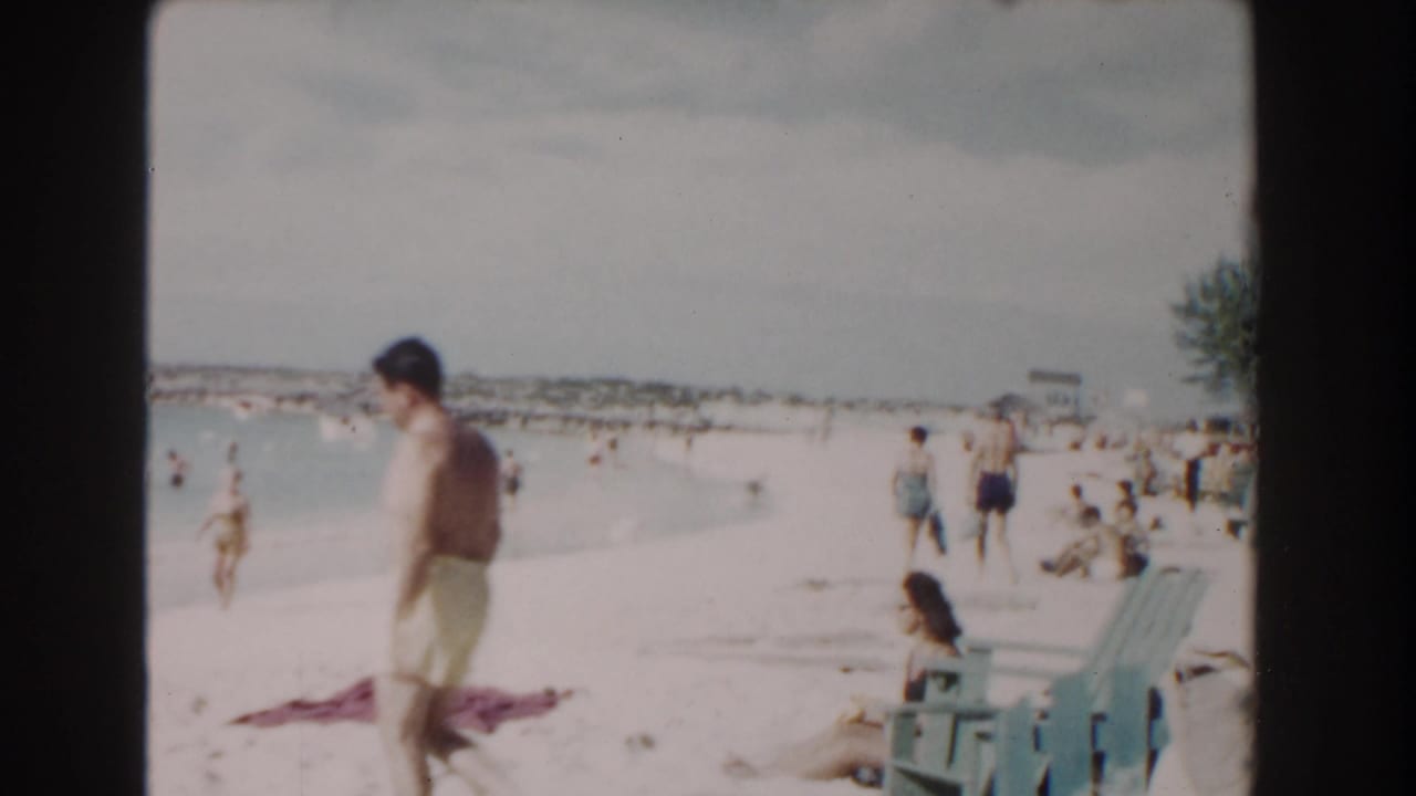 Man in yellow swim trunks walks along a crowded beach beside calm blue water.