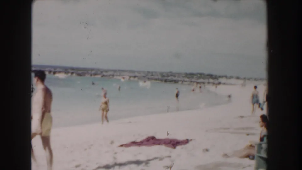1961 Nassau beach footage showing a shoreline with swimmers, walkers, a seated person, and a towel on the pale sand.