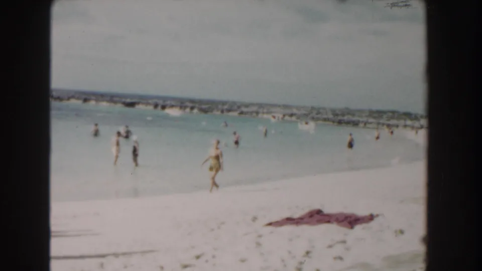 Nassau beach scene with people wading in calm water, a figure walking on sand, and a towel in the foreground.