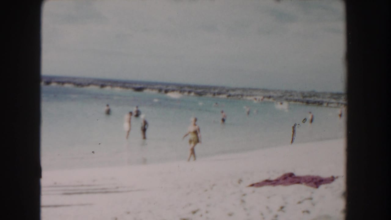 Bathers wade and stand in shallow water beside a pale sandy beach.
