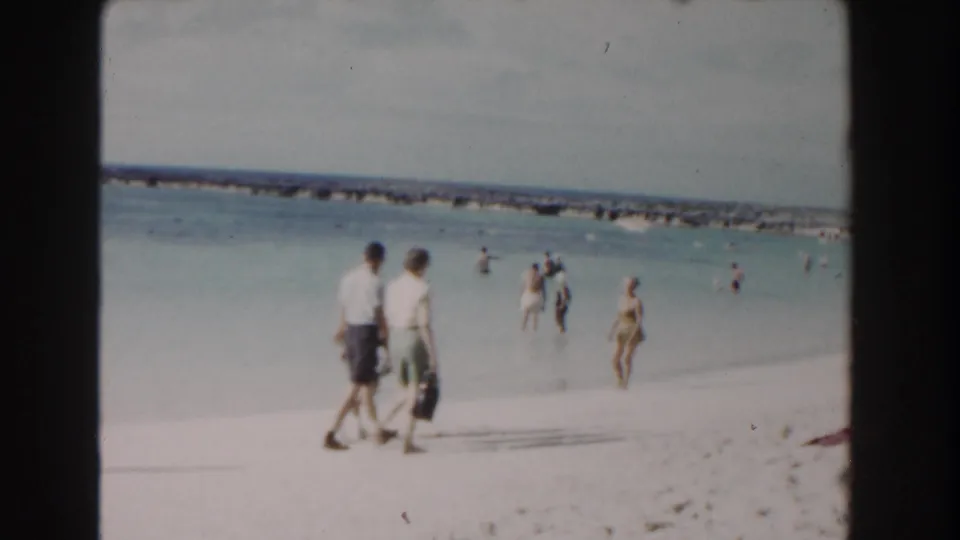 Two beachgoers stroll across a Nassau shoreline as swimmers and waders move through the shallow water in the background.