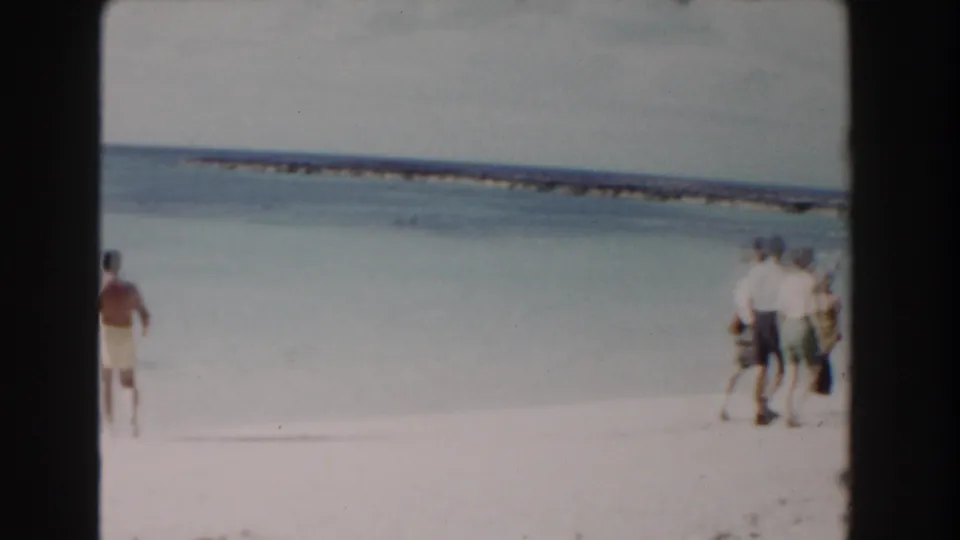 Wide 8mm view of a Nassau beach, with a person moving near the surf and small groups standing along the waterline.