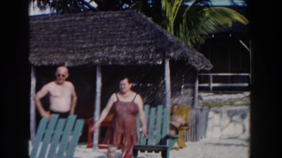 Two older adults near blue beach chairs and a thatched shelter, with palm fronds and resort-like beach structures behind them.