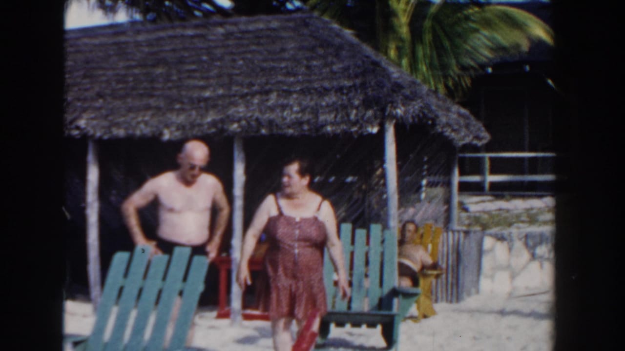 A couple walks on a sandy beach near a thatched cabana and painted chairs.