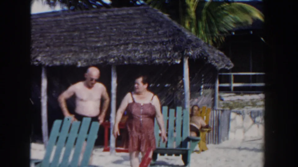 In Nassau, Bahamas, 1961, a shirtless man and a woman in a red swimsuit walk past beach chairs and a thatched cabana.