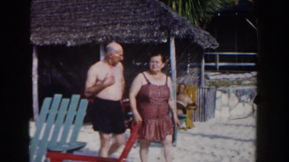 The man and woman walk closer on a Nassau beach, framed by painted chairs, a thatched shelter, and another reclining tourist.