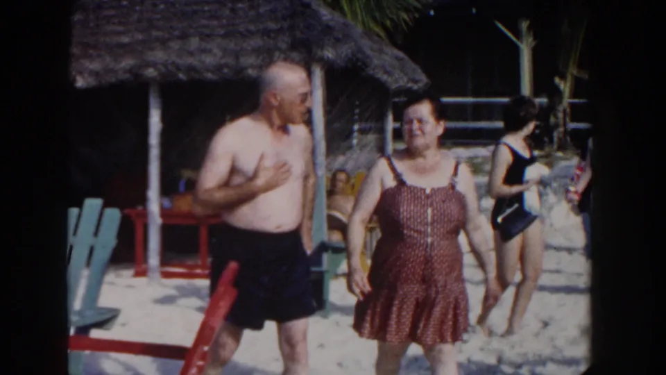The couple continues toward the camera on the Nassau beach while a woman in a dark swimsuit crosses behind them near the cabana.