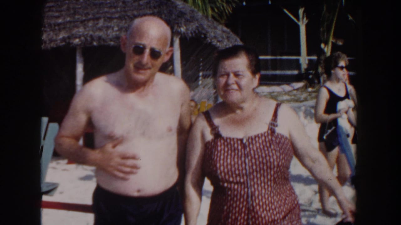 Older couple posing together on a sunny beach near a thatched resort shade structure.