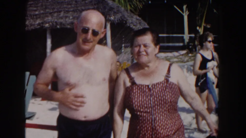 In Nassau, Bahamas, 1961, an older couple poses for a home-movie camera beside a sandy resort area with chairs and thatched roofing.