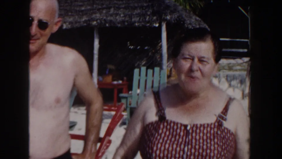 The couple remains near the Nassau beach setting, with the man in sunglasses and the woman in a patterned swimsuit framed by palm shade and resort seating.