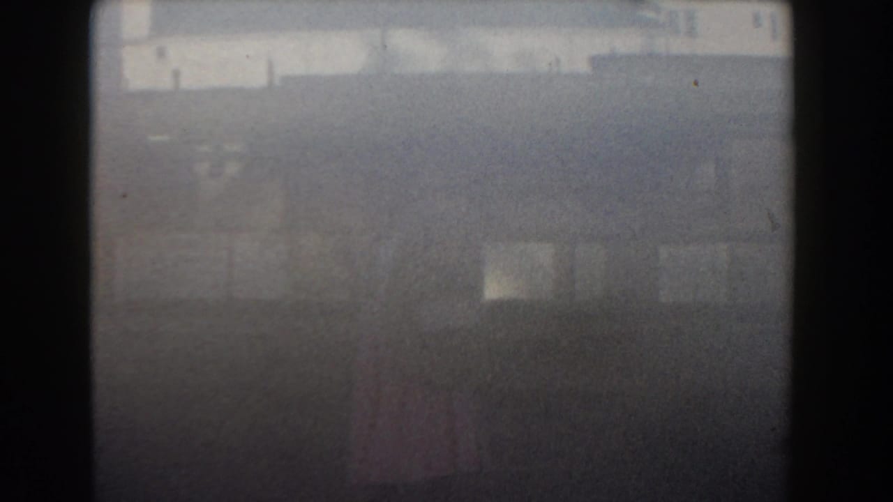 Soft 1961 Nassau home-movie frame showing a distant woman in pink near pale buildings under hazy daylight.