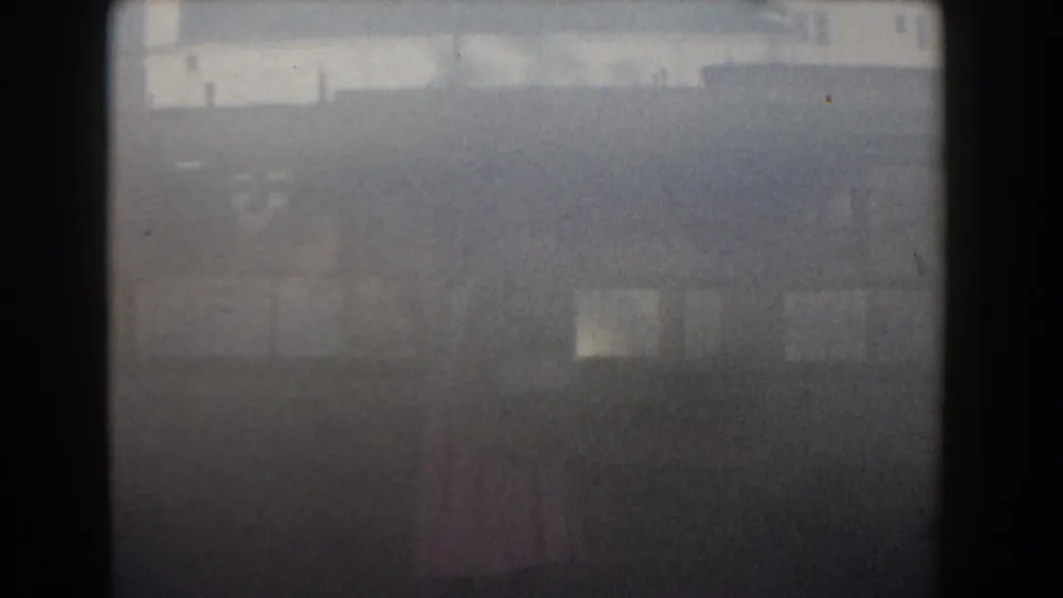 Soft 1961 Nassau home-movie frame showing a distant woman in pink near pale buildings under hazy daylight.