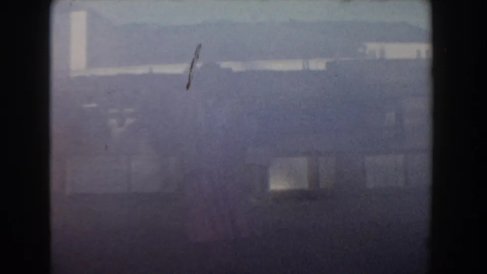 Vintage 8mm frame from Nassau with a distant pink-dressed figure, pale building shapes, and strong haze.
