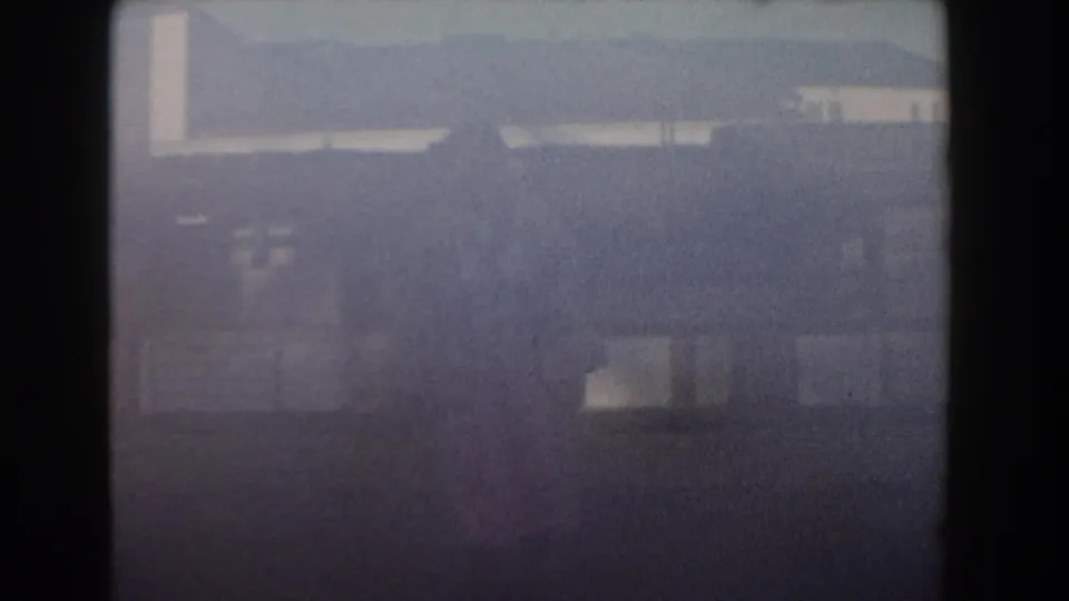 Soft 1961 Nassau home-movie frame with pale building forms, purple-blue tint, and a faint distant woman.