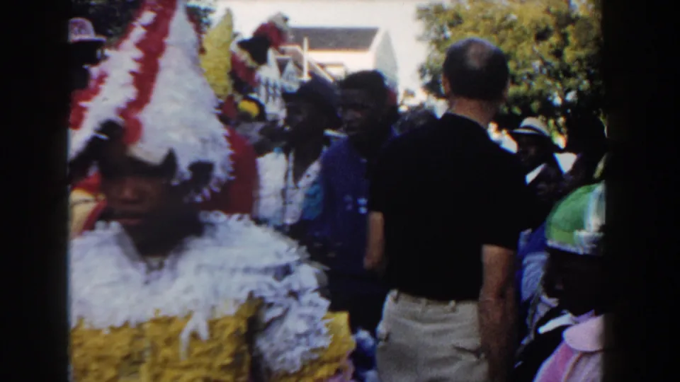 A costumed marcher with a red and white feathered headdress moves through a Nassau street crowd as spectators stand nearby.