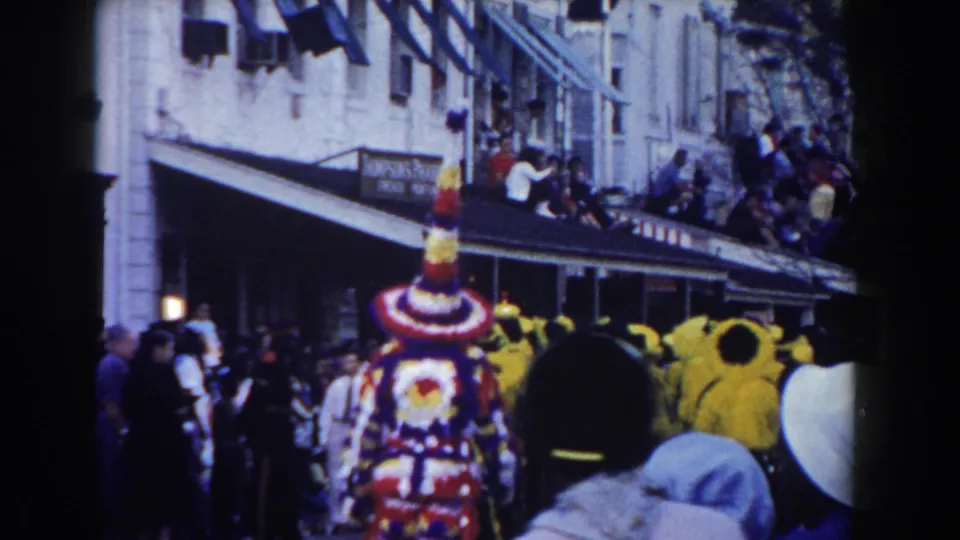 A tall decorated costume and yellow-clad performers appear on a Nassau street as spectators watch from sidewalks and balconies.