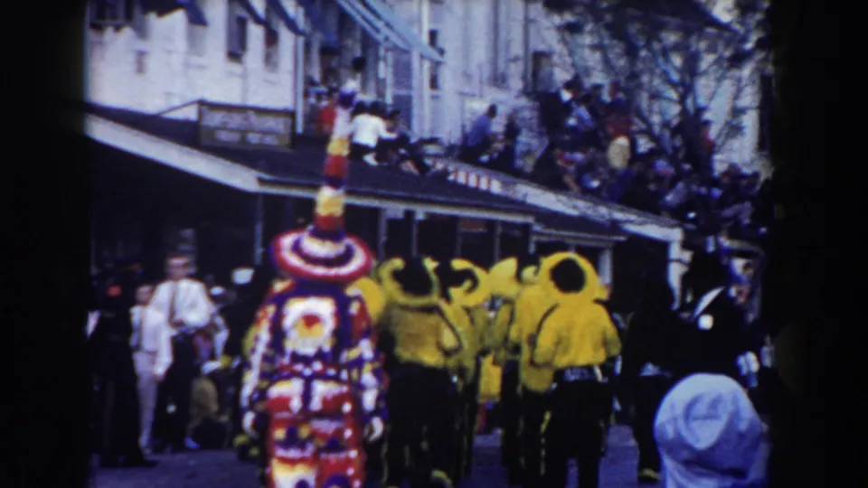 Yellow-clad parade performers gather around a tall decorated costume in Nassau, with spectators lining the street and nearby buildings.