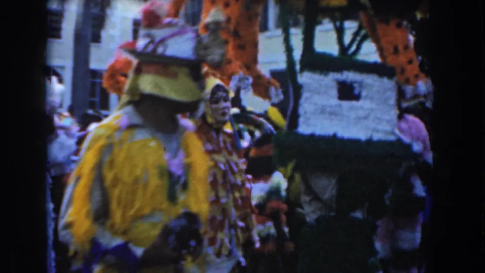 1961 Nassau parade frame with a yellow-costumed marcher, masked participant, and large handmade decoration in the street.