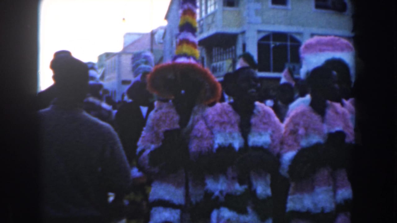 Costumed marchers in a 1961 Nassau parade pass near the handheld camera, with spectators and buildings behind them.