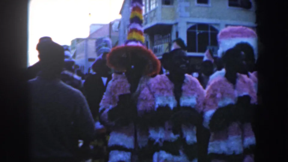 Costumed marchers in a 1961 Nassau parade pass near the handheld camera, with spectators and buildings behind them.