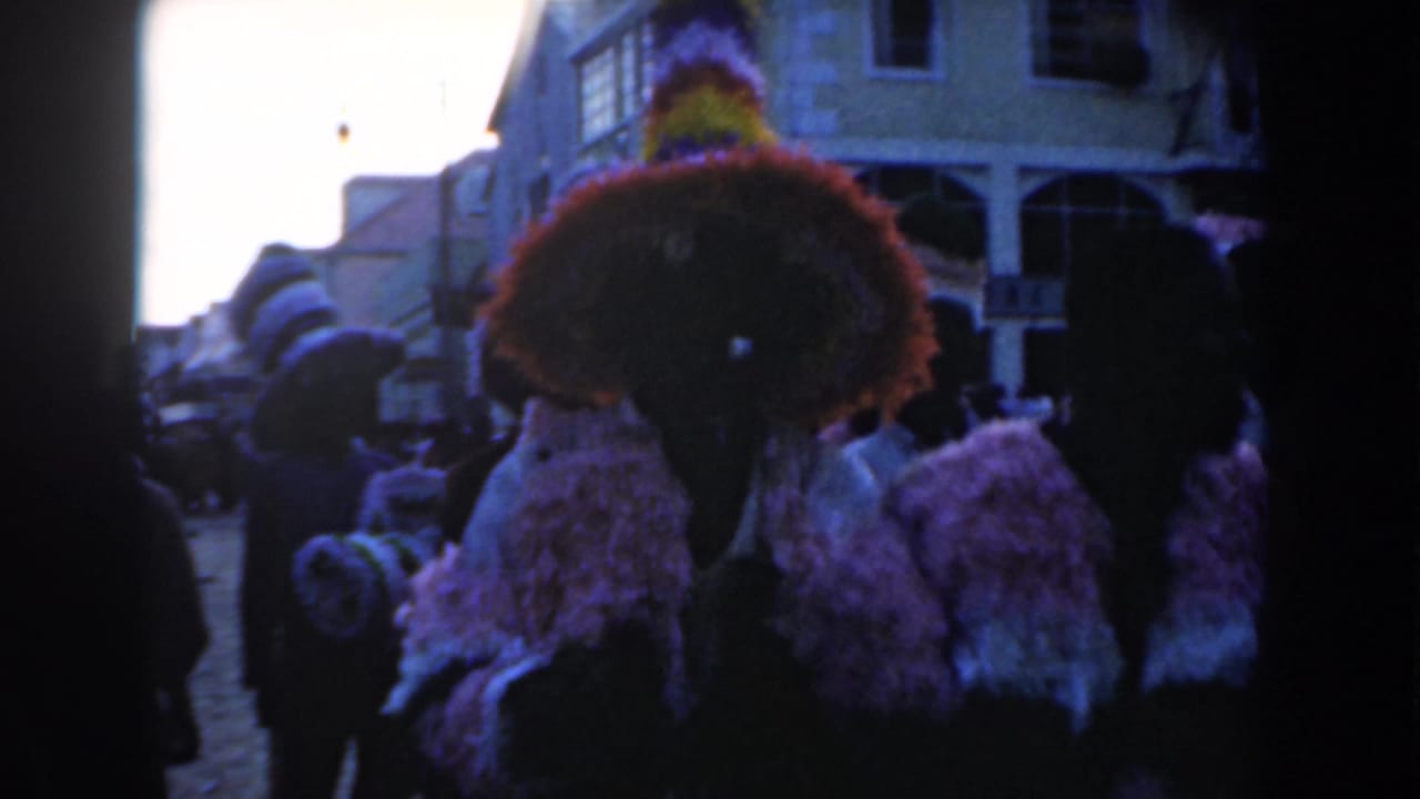 A close handheld view shows a large feathered headpiece and striped costume during the Nassau parade, with the street receding behind.