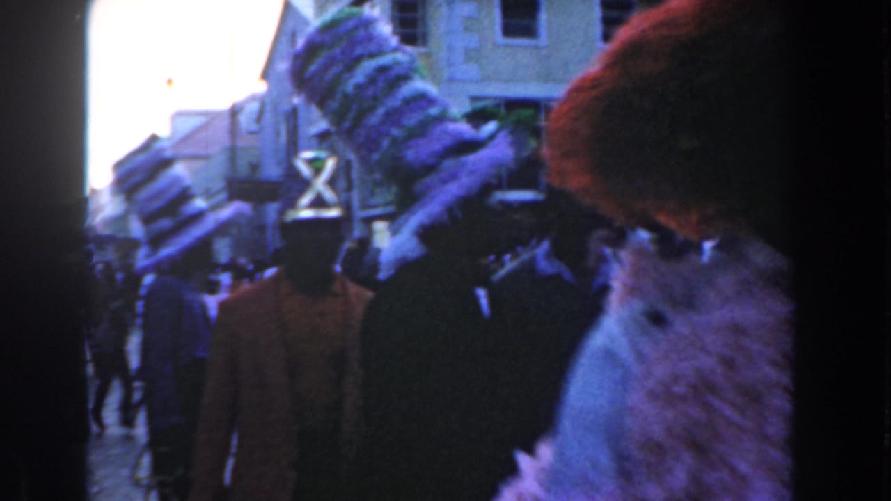 1961 Nassau parade participants with tall feathered headpieces move through a crowded street, partly obscuring the lens.