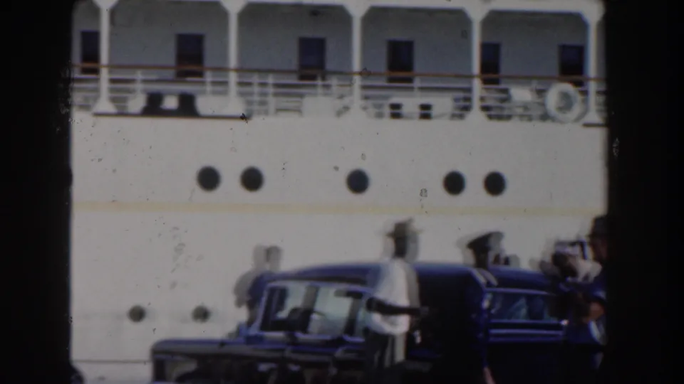 Port-side view in Nassau with men in light clothing and hats near dark sedans below the portholes of the cruise ship.