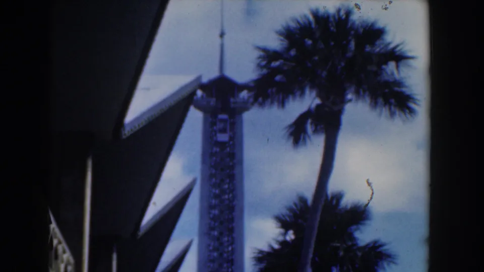 1961 Nassau travel frame with a palm crown crossing an observation tower near dark angular building overhangs.
