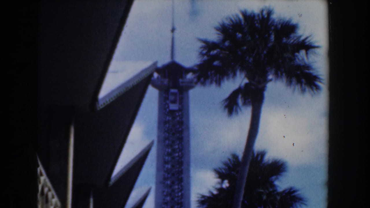1961 Nassau home-movie frame showing a white tower, palm trees, blue sky, and angular foreground architecture.