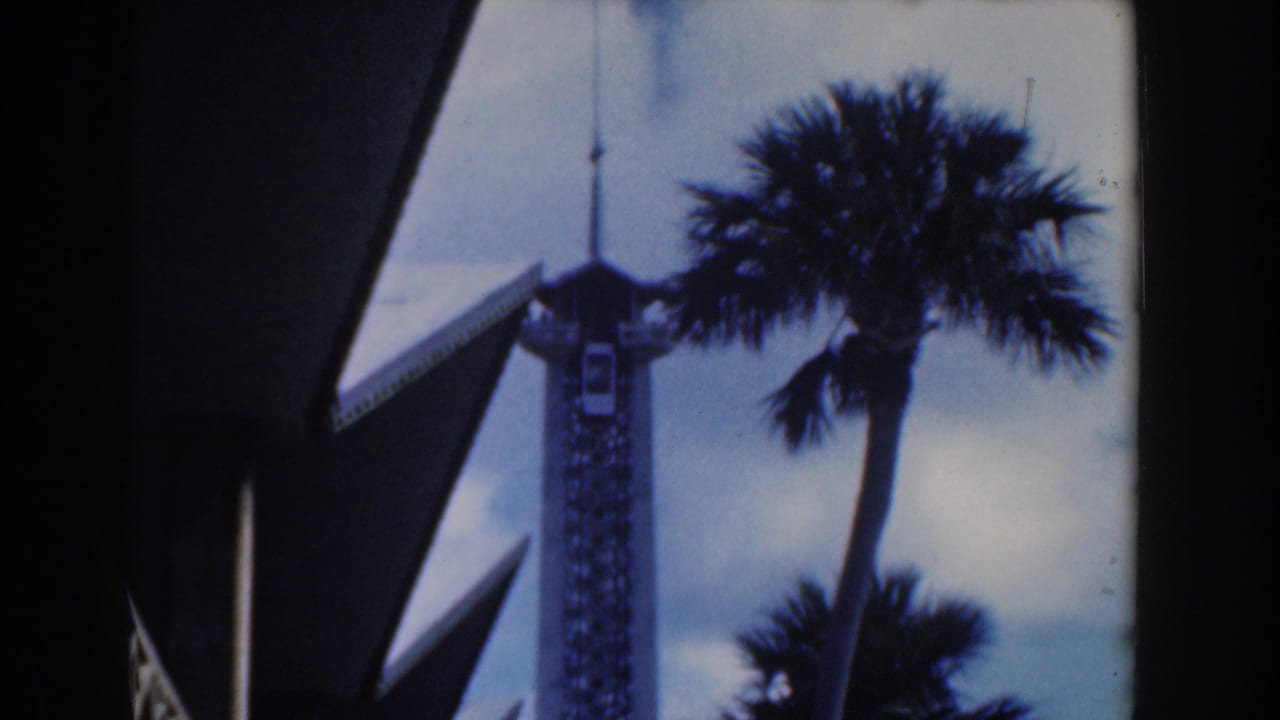 Palm tree and white tower against a pale blue sky near angular architecture.