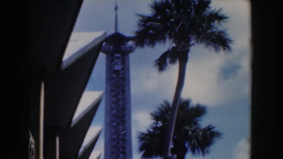 1961 Nassau home-movie frame showing a tall spired tower behind palm trees and modern rooflines.