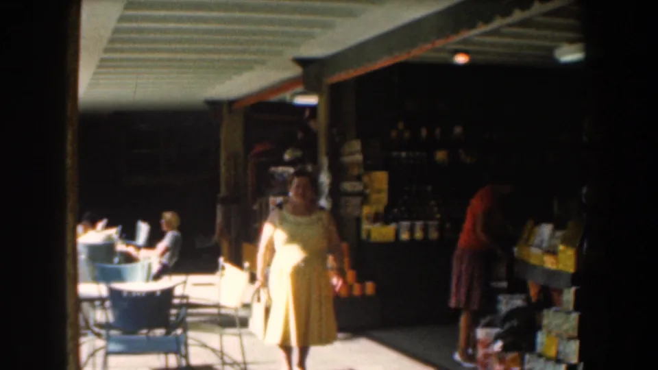 1961 Nassau patio restaurant scene with a woman in a yellow dress, seated people, and goods under a covered area.