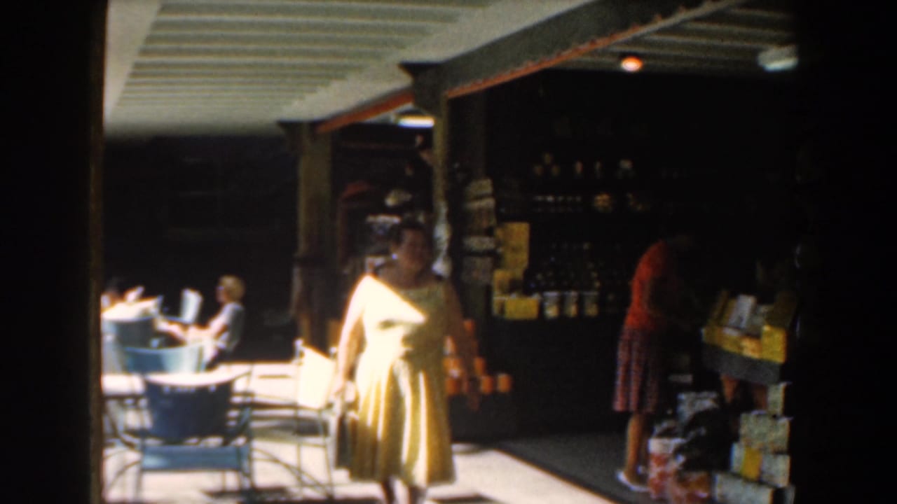In Nassau, Bahamas in 1961, a woman in a yellow dress walks through a shaded open-air corridor near seated people and merchandise shelves.