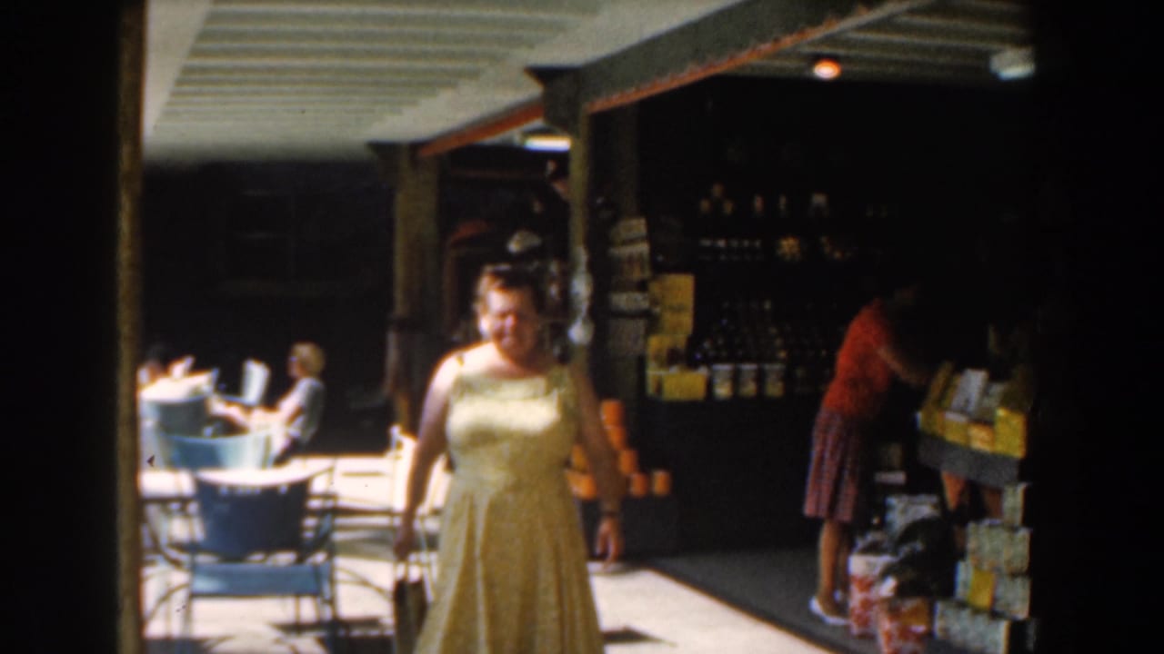 Woman in a yellow dress nears the camera while another person stands by shop shelves.