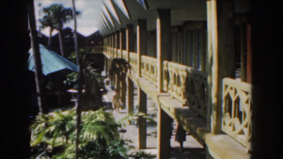 A Nassau 1961 exterior shows wooden railings, columns, palm plants, and a sunlit walkway beside the tropical building.